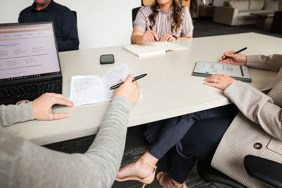 4 employees work together to discuss market research for a client, making notes and working on their computer